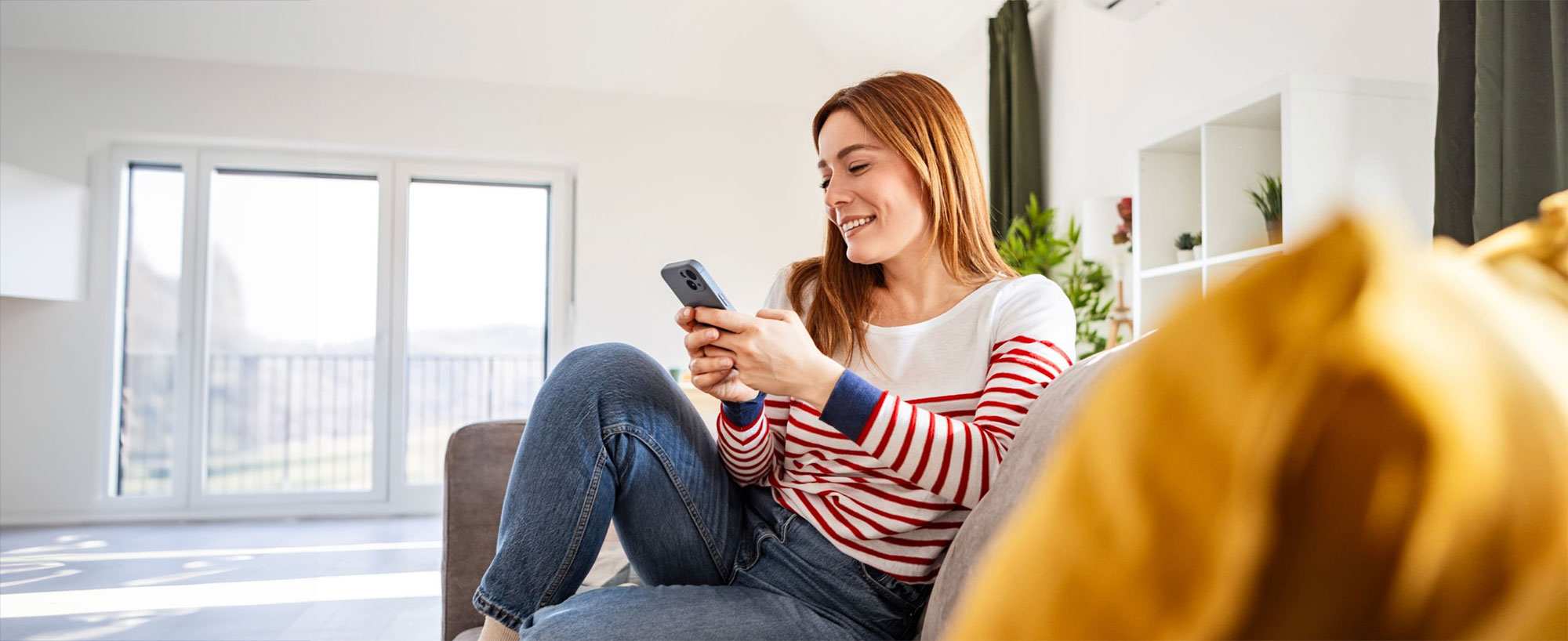 Mujer distendida en sofa de sala de estar sonriendo mientras utiliza su celular