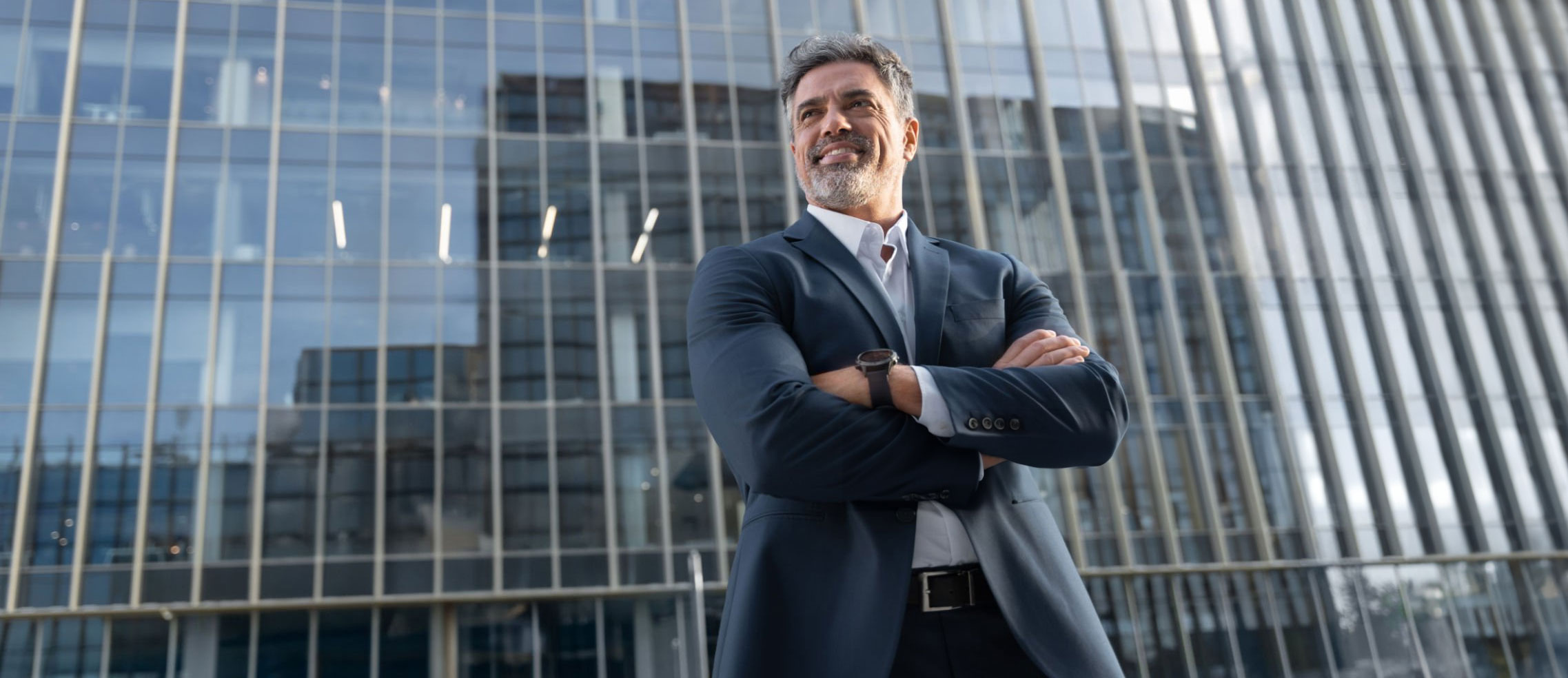 Hombre adulto de traje sonriendo seguro y confiado mirando al horizonte con edificio de ventanas translúcidas como fondo