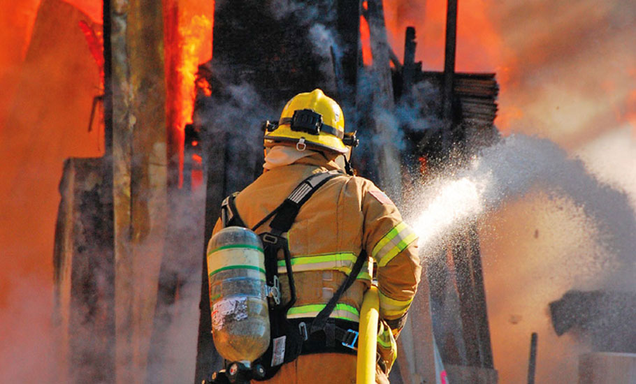 Bombero bien equipado de espaldas intentando sofocar gran incendio con manguera